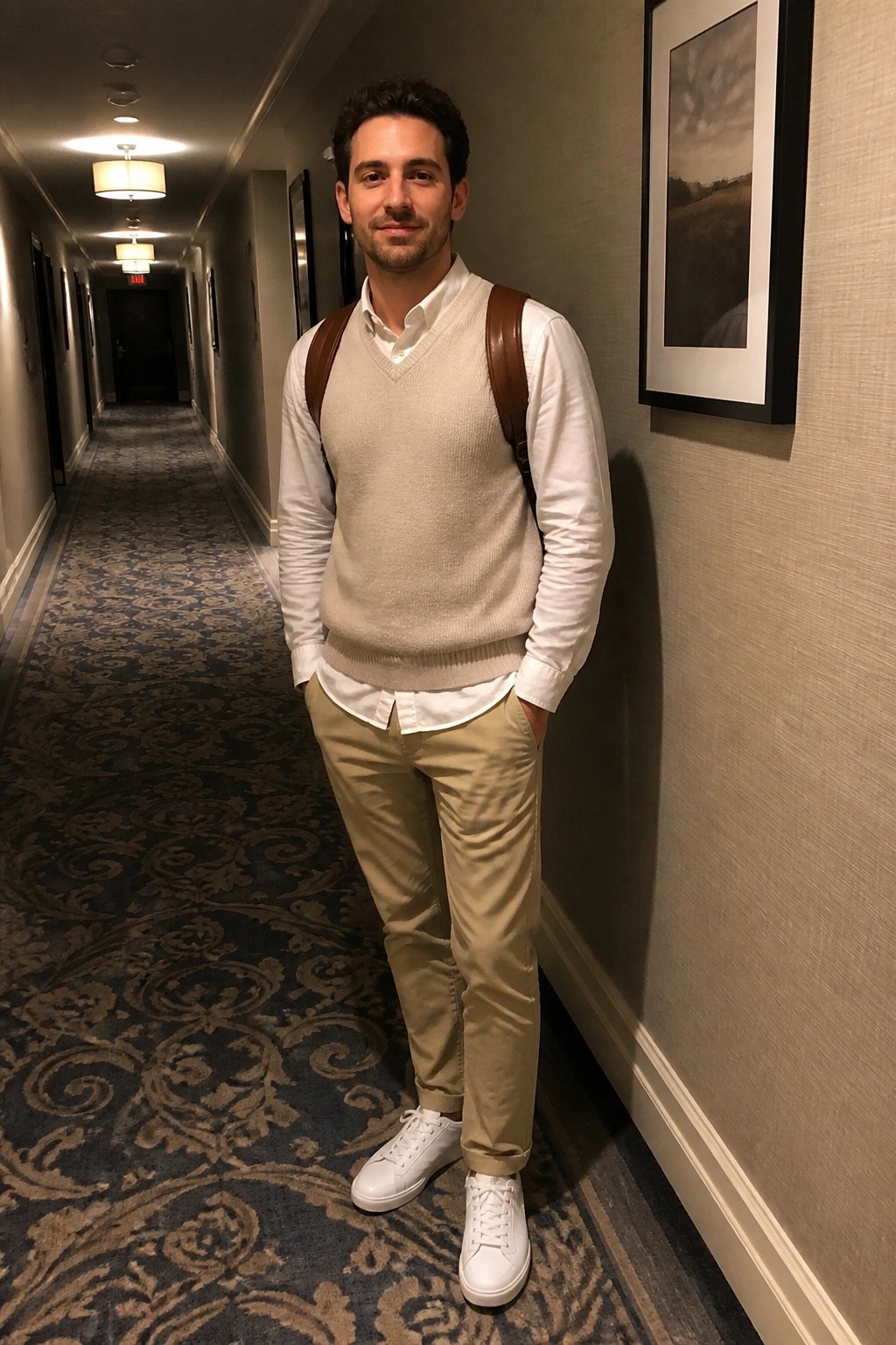 Man in cream sweater vest and khaki pants with backpack in hotel hallway.