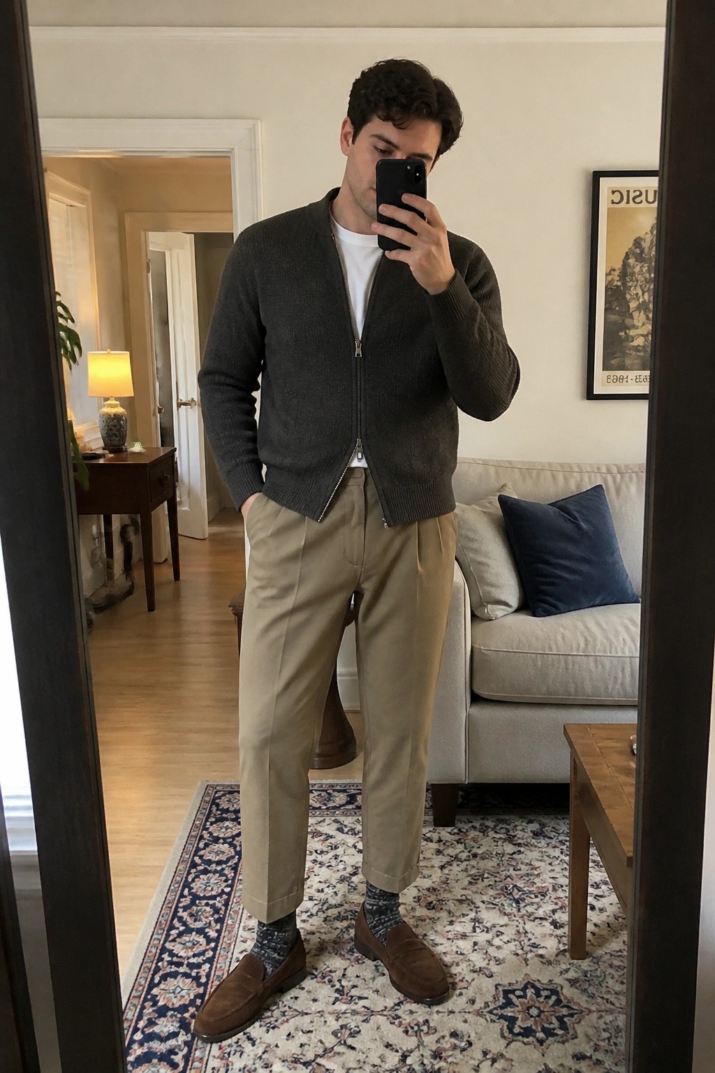Man in mirror selfie wearing dark gray zip-up cardigan over white t-shirt with khaki pants, brown suede loafers, and patterned socks in a living room setting.