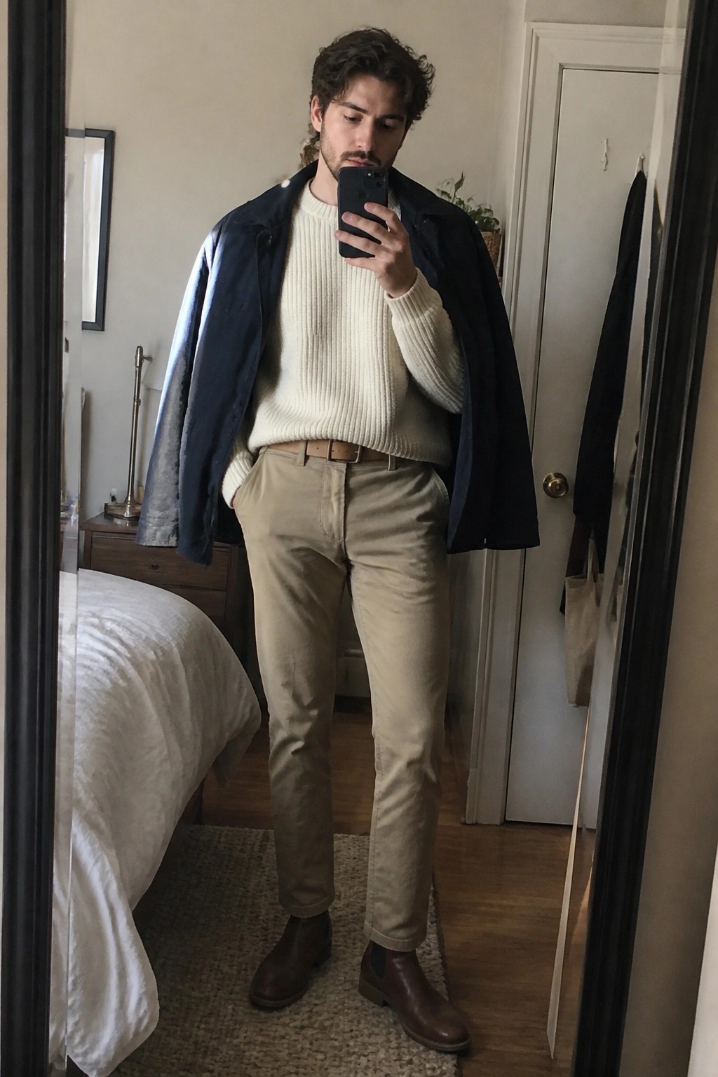 Man in cream sweater, navy jacket, khaki pants, and brown boots taking mirror selfie.
