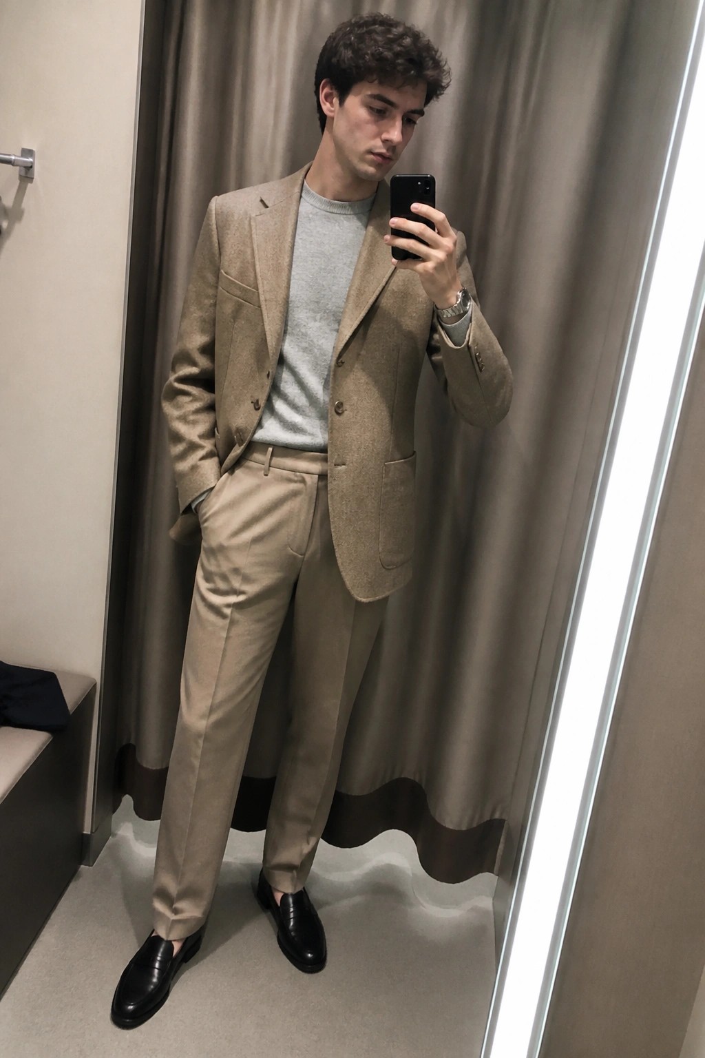 Man in beige suit and gray sweater taking mirror selfie in fitting room