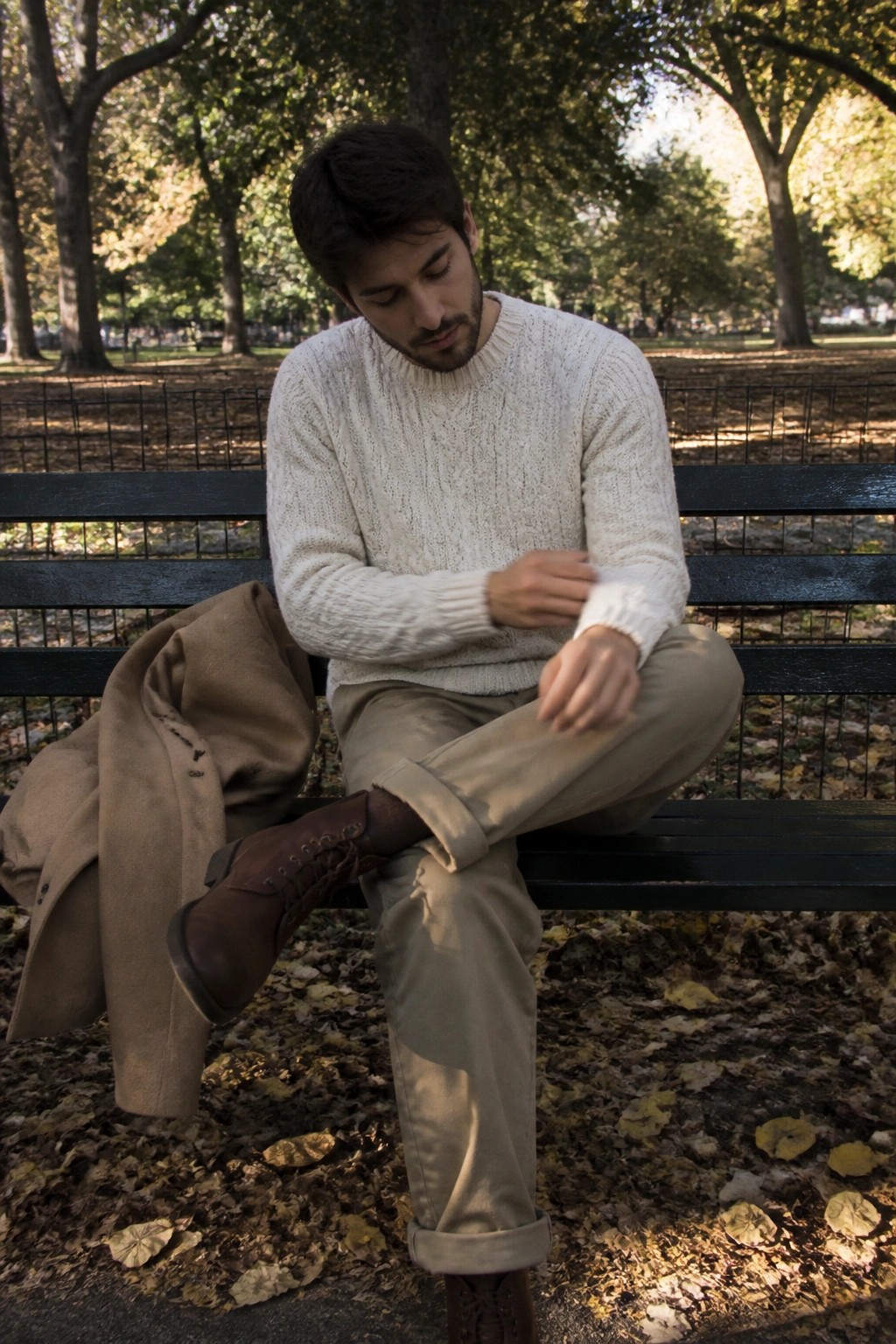Man in white sweater on park bench beside tan coat amid autumn leaves