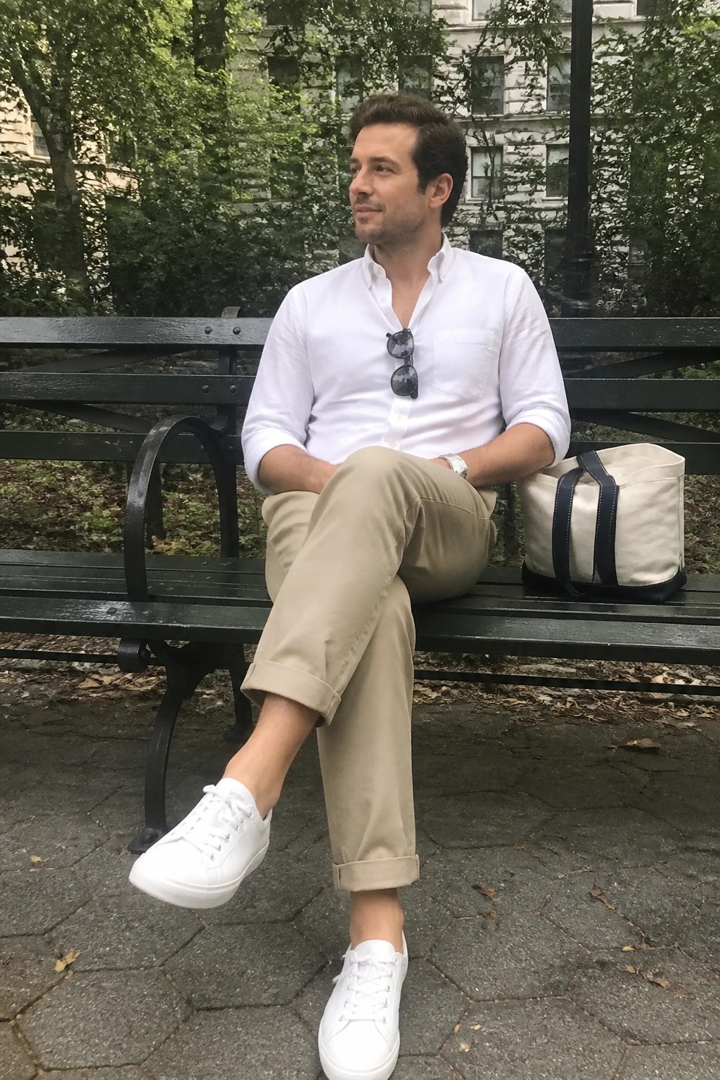 Man in white shirt and khaki pants on green park bench with tote bag