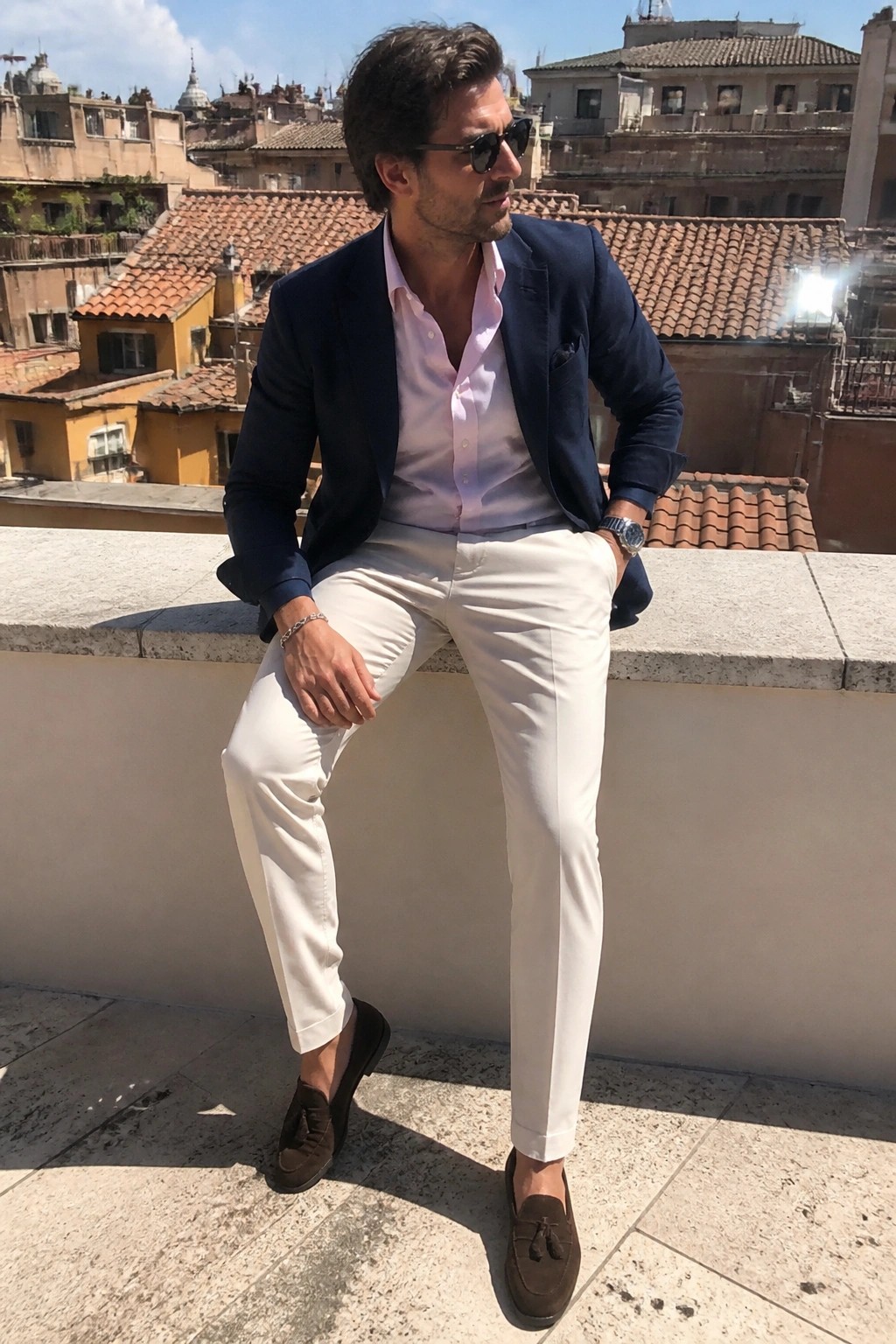 Man in navy blazer and white pants seated on ledge overlooking city rooftops