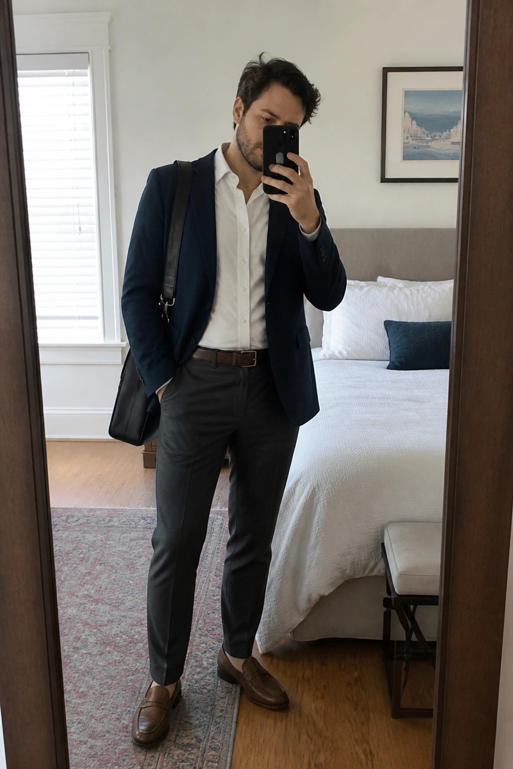 Man in navy blazer and white shirt taking mirror selfie in bedroom