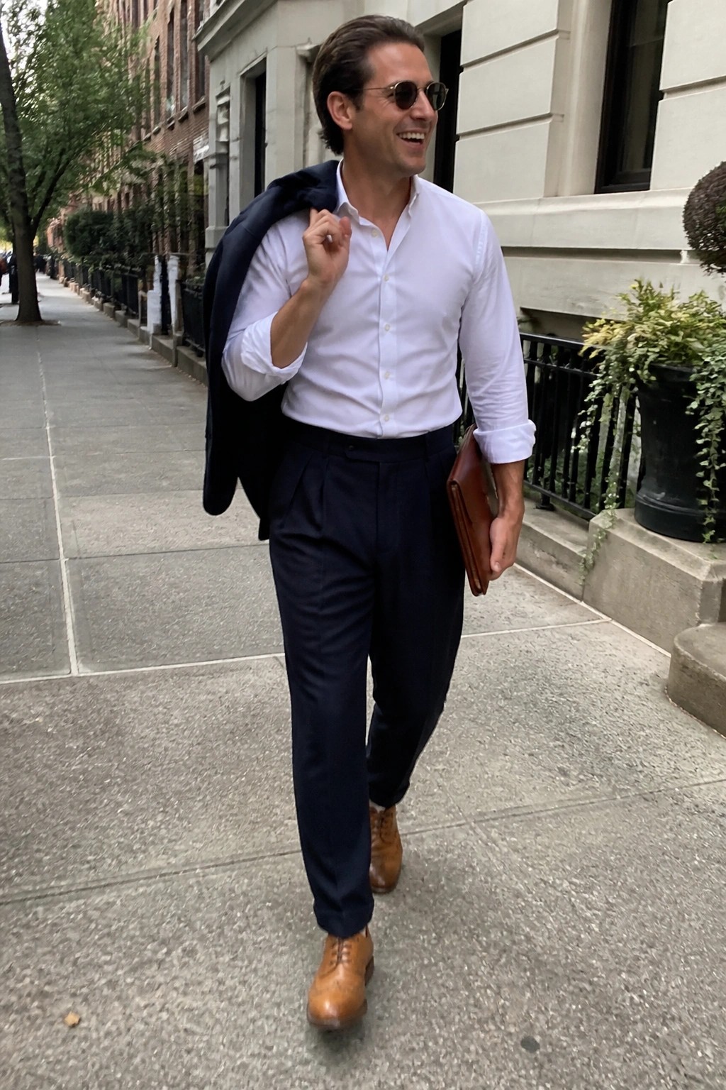 Smiling man in white shirt carrying navy blazer and leather folder on city sidewalk.
