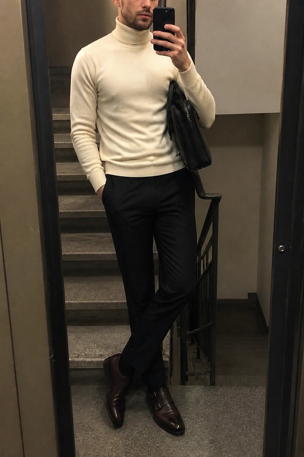 Man in cream turtleneck, black pants, and brown monk shoes taking mirror selfie