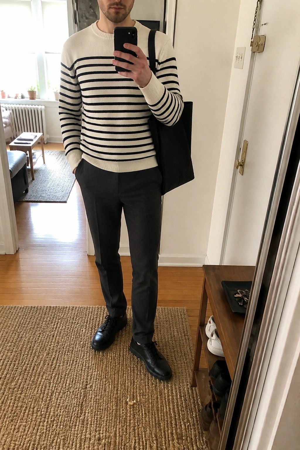 Man in striped sweater and dark pants taking mirror selfie with tote bag.
