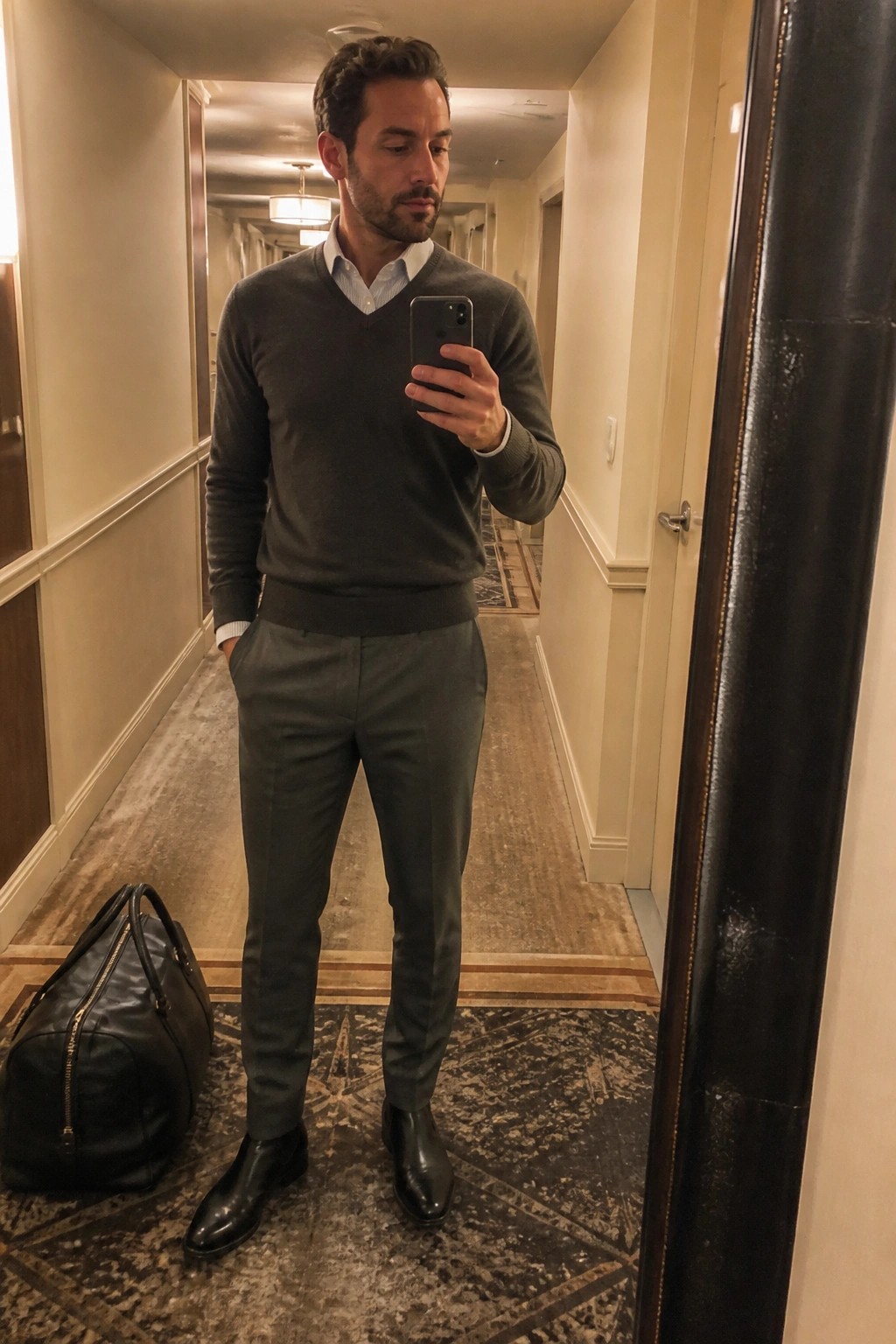 Man in gray sweater and trousers taking mirror selfie beside black duffel bag in hallway