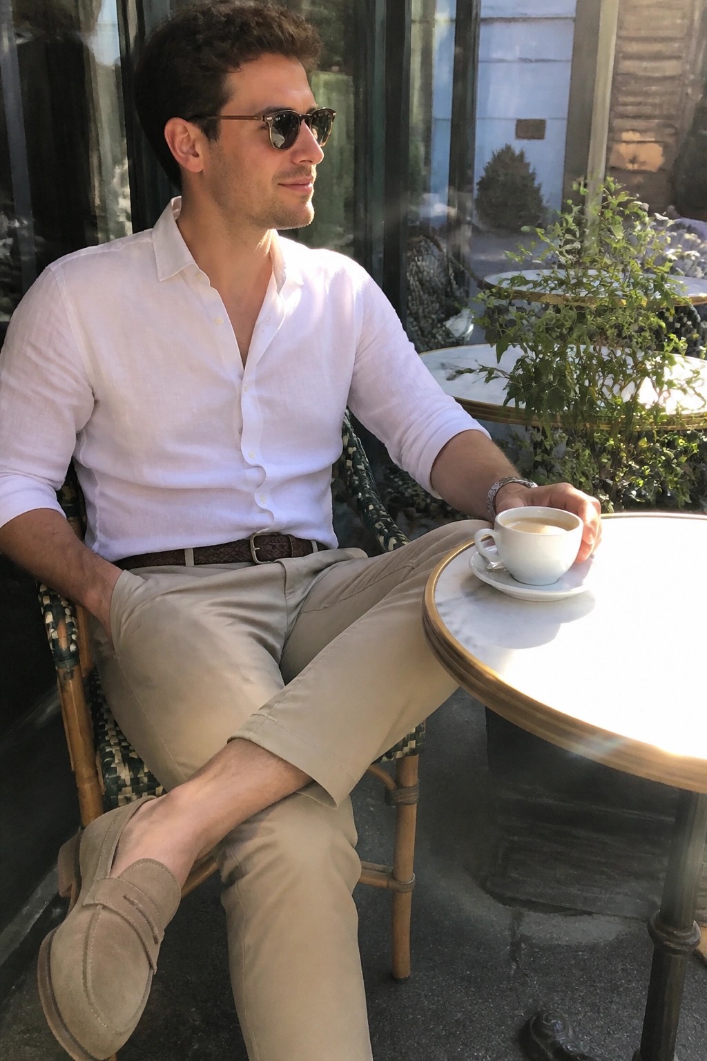 Man in white shirt and beige pants at sunny outdoor café table with coffee.