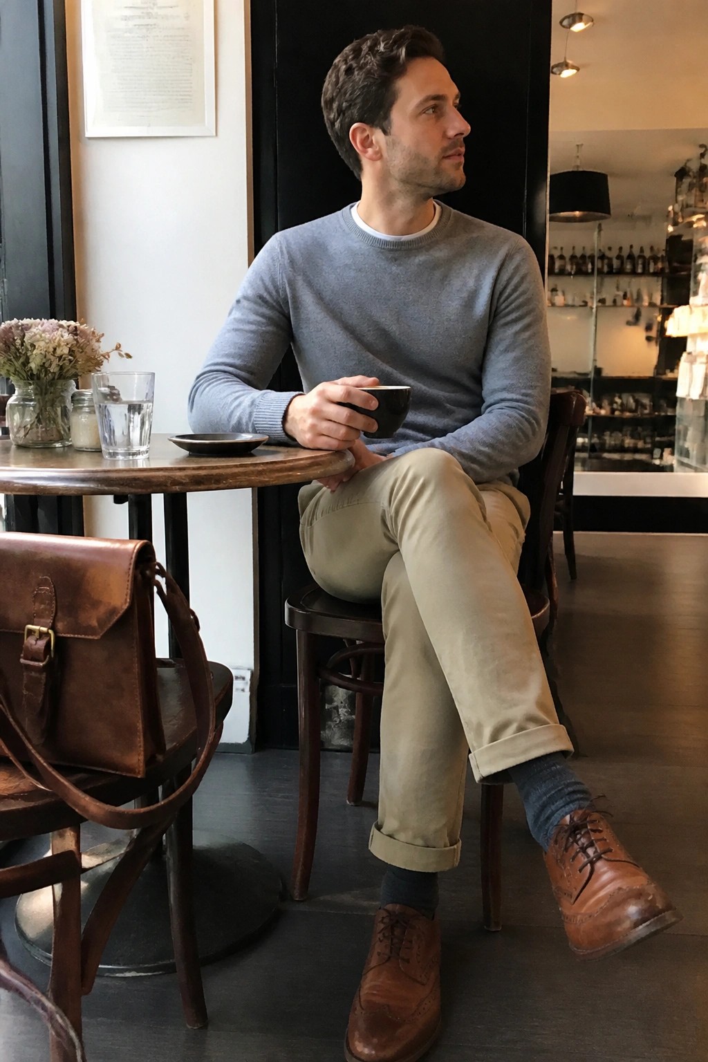 Man in gray sweater and beige pants sits in cafe holding black cup, leather bag nearby