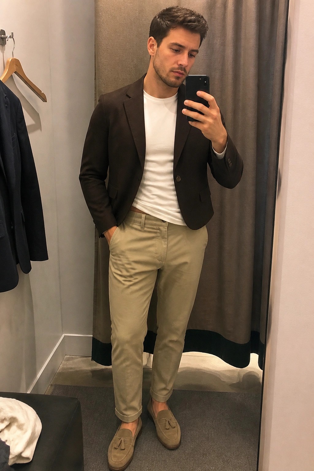 Man in brown blazer and khaki pants taking mirror selfie in fitting room
