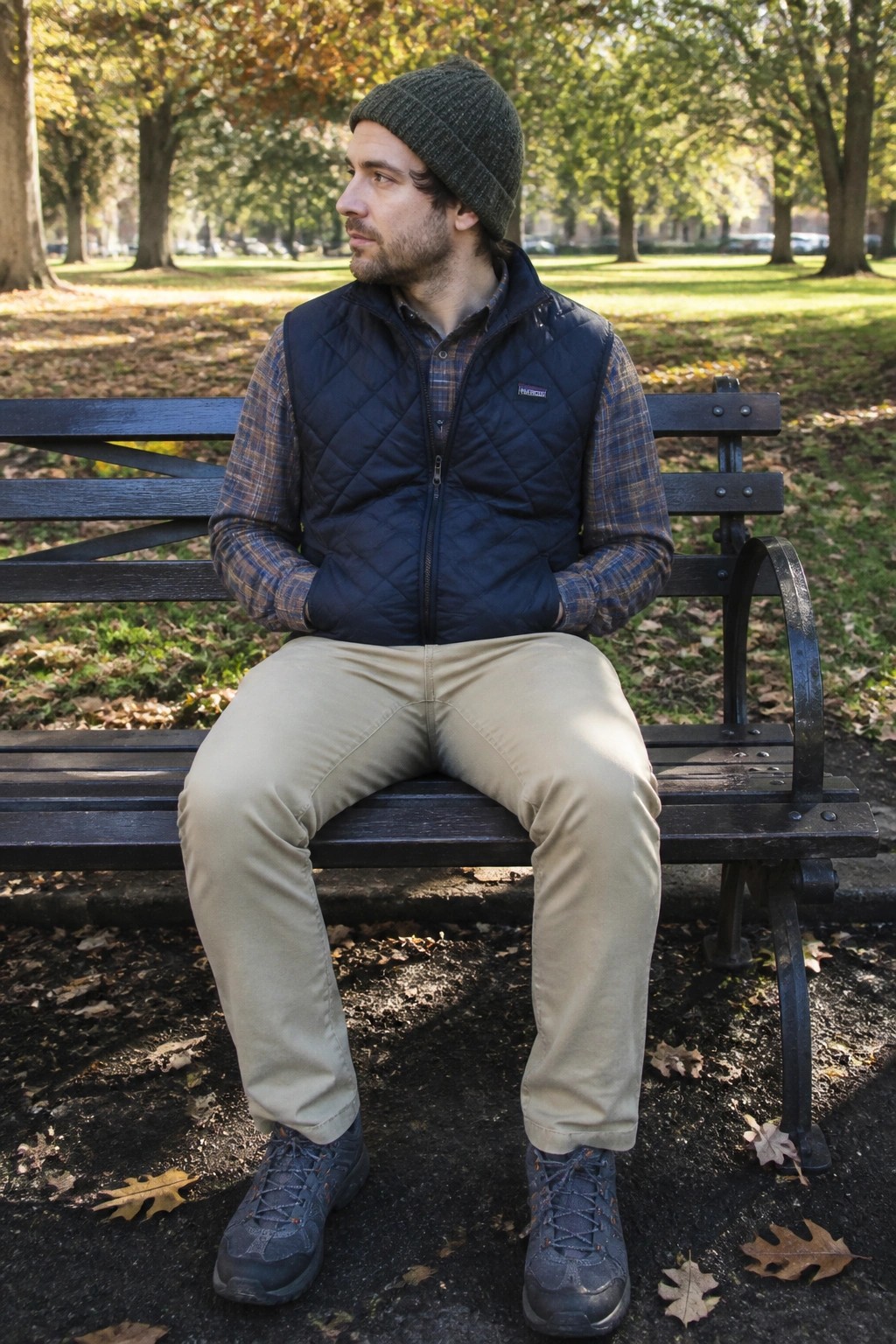 Man in green beanie and blue vest sitting on park bench amid autumn leaves.