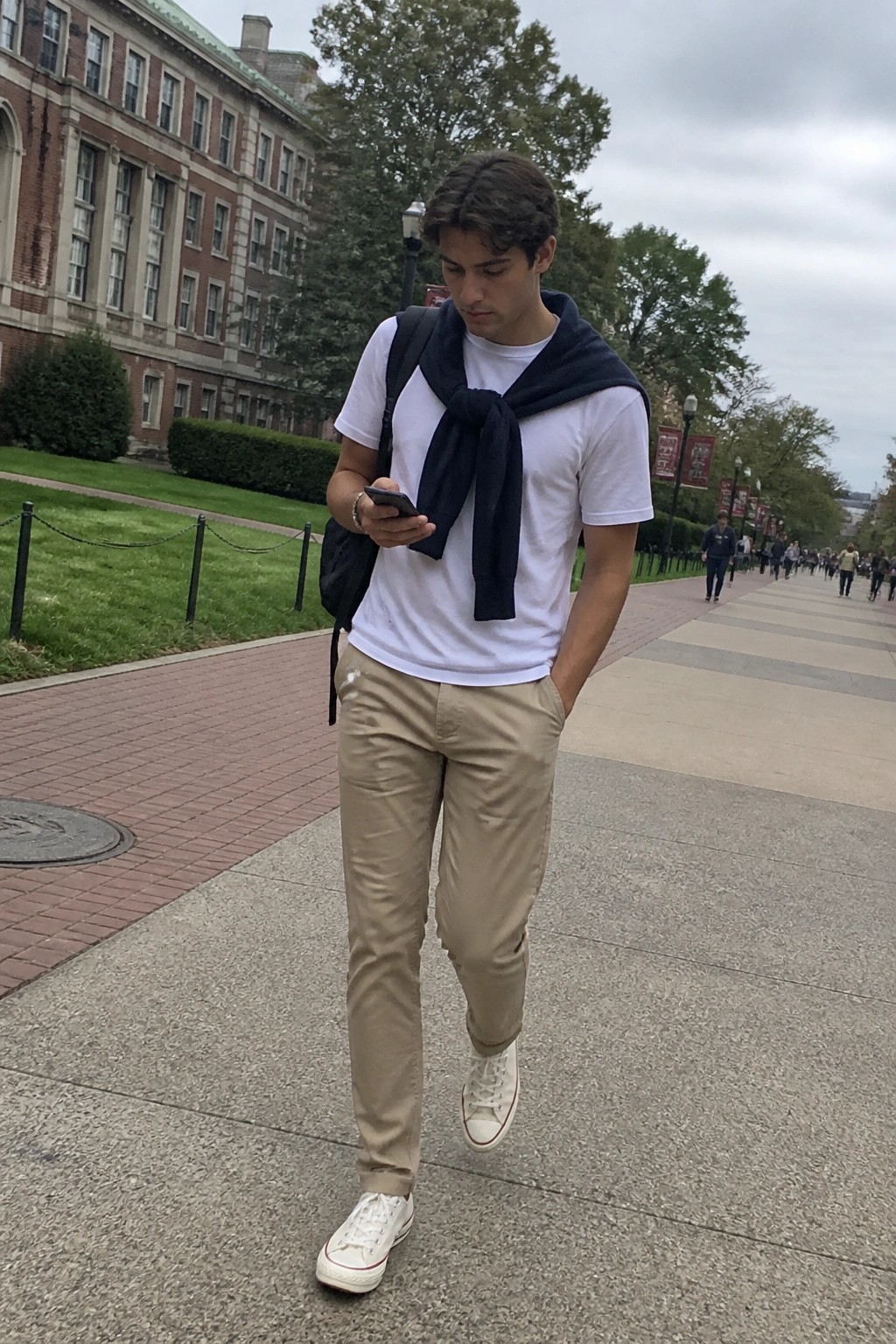 Man wearing beige khaki chinos, white crew neck t-shirt, navy sweater tied at the neck, white low-top sneakers, and carrying a black backpack while looking at a phone.