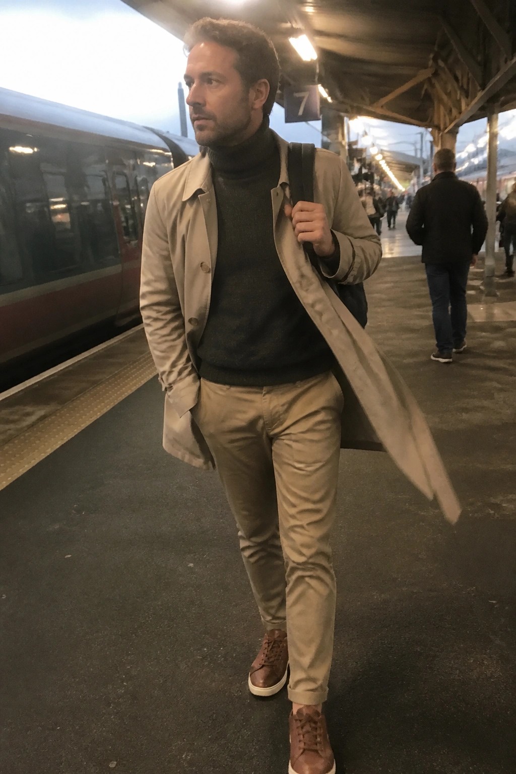Man in beige trench coat and turtleneck on train station platform.