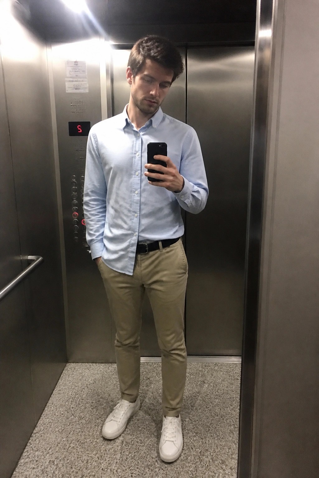Man wearing a light blue button down shirt tucked into khaki chinos with a black belt and white sneakers while holding a phone in an elevator.