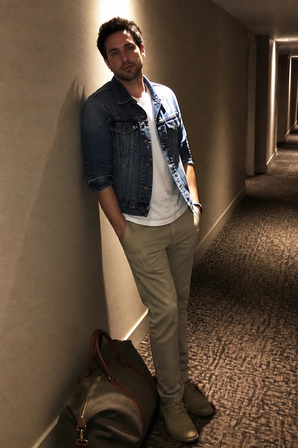 Man in denim jacket leaning against hallway wall beside olive duffel bag