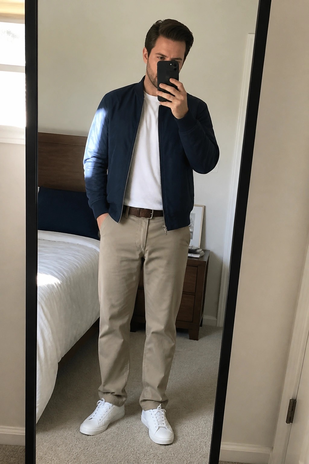 Man in navy bomber jacket, white tee, khaki pants taking bedroom mirror selfie