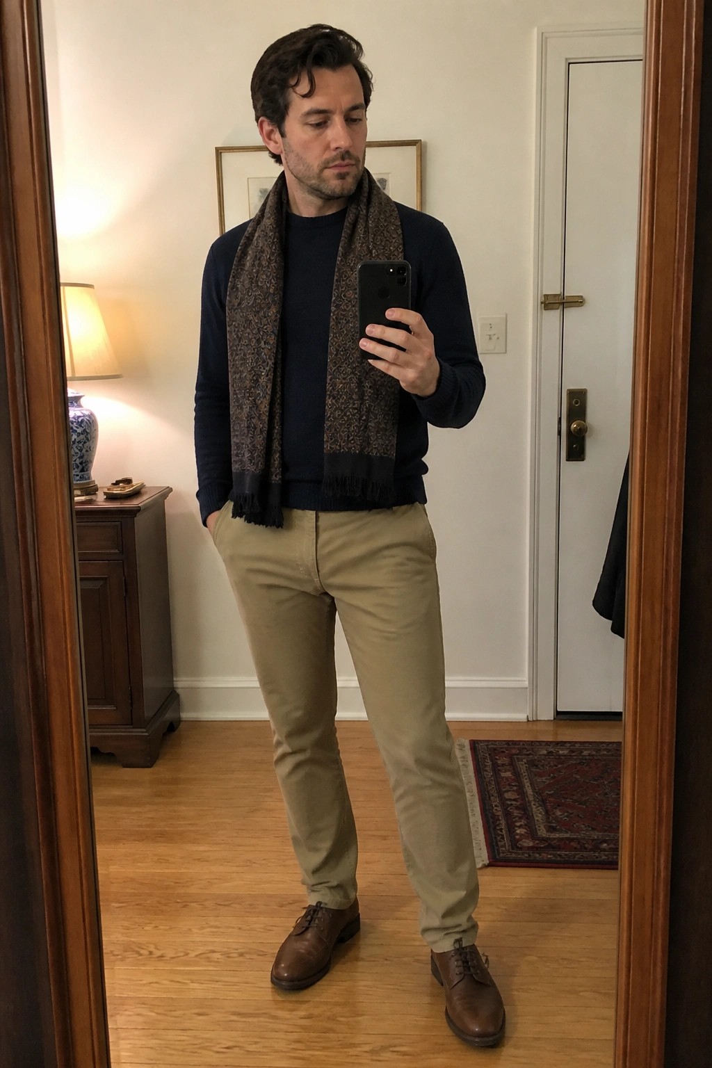 Man in navy sweater and patterned scarf taking mirror selfie in entryway.
