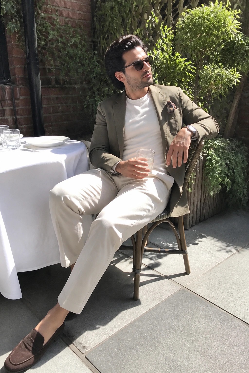 Man in olive blazer and beige pants holding glass on sunny patio