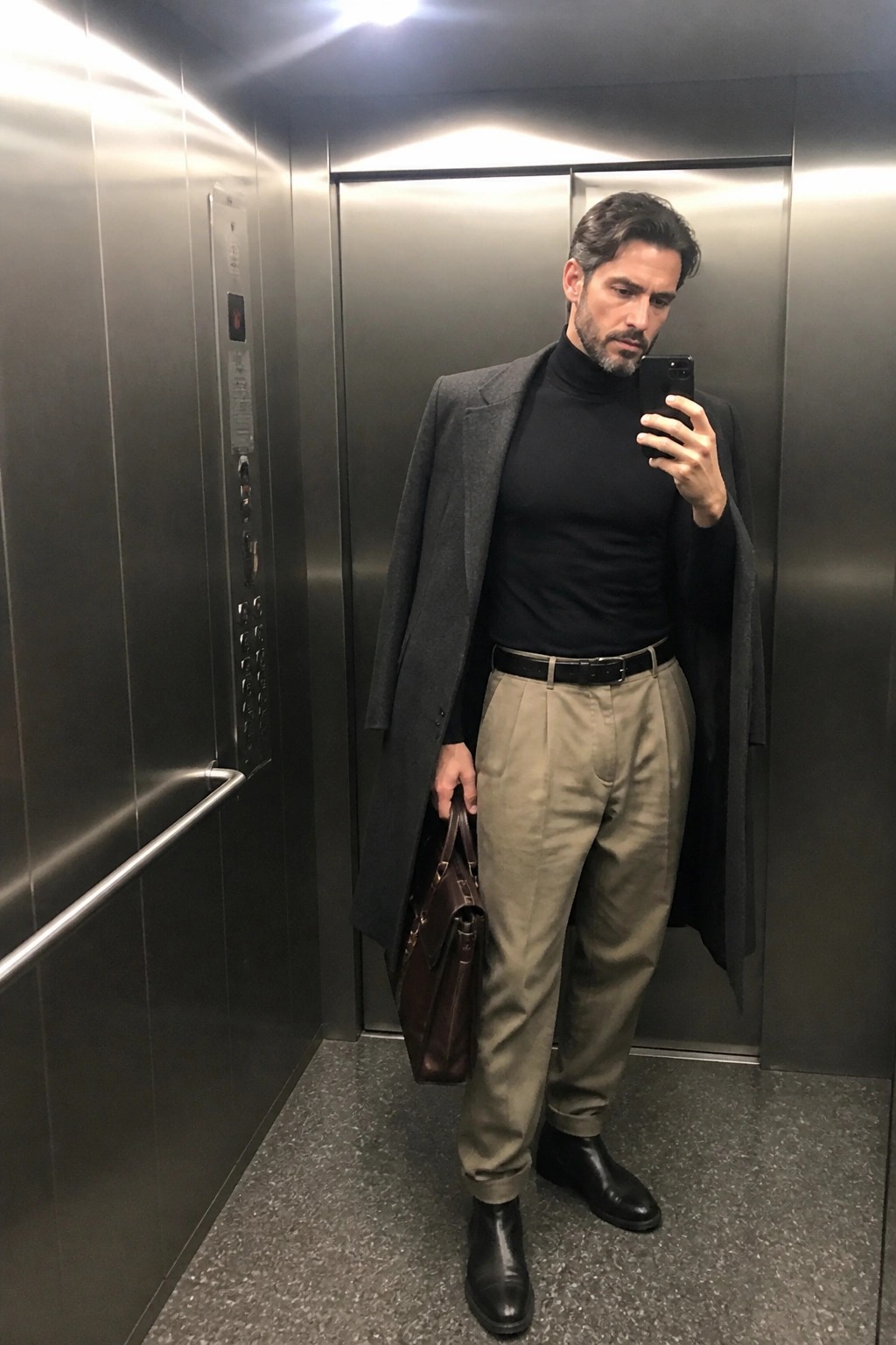 Man standing in elevator wearing relaxed fit khaki pants, black turtleneck, gray wool coat, black Chelsea boots and holding a brown leather bag.