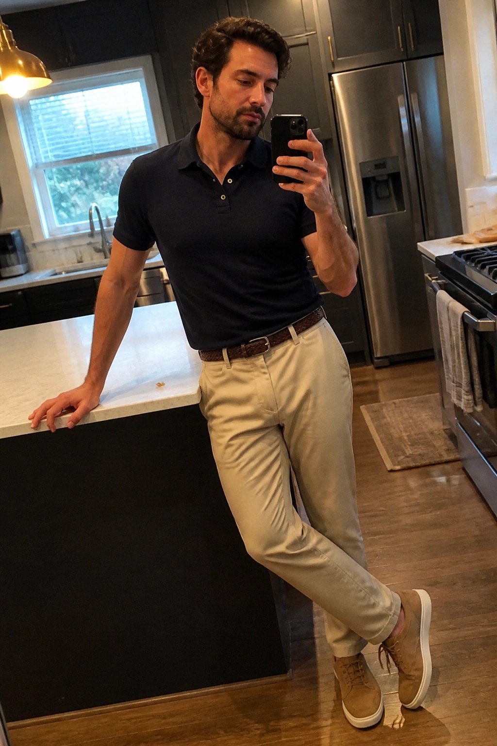 Man in navy polo and beige pants taking mirror selfie in kitchen