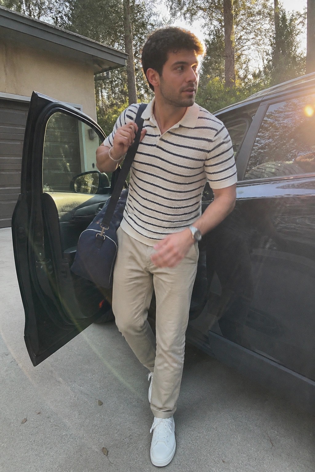 Man wearing a cream and navy striped polo shirt tucked into relaxed fit khaki pants with white sneakers and a navy bag slung over one shoulder.