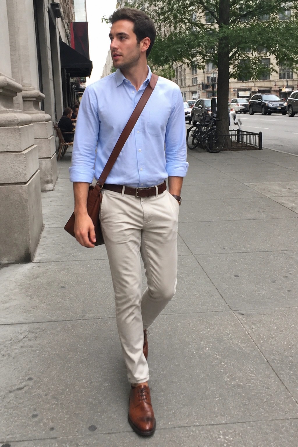 Man in light blue shirt and beige pants with brown leather bag on sidewalk