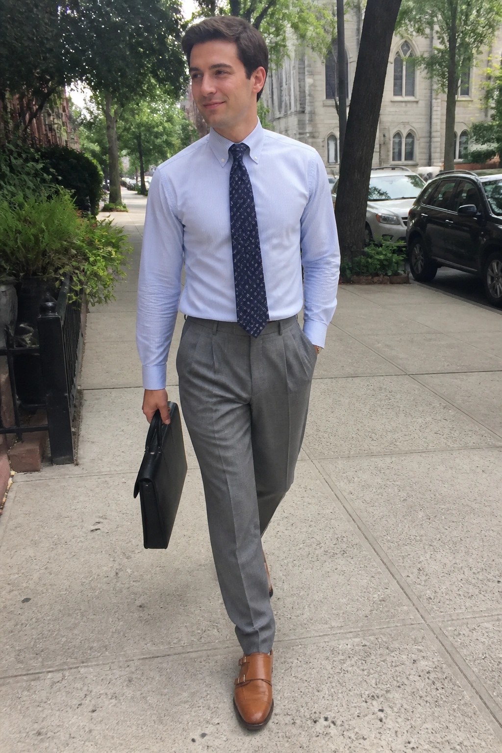 Man wearing light blue button down shirt, navy patterned tie, gray pleated trousers, brown double monk strap shoes while holding a black briefcase on a city sidewalk.