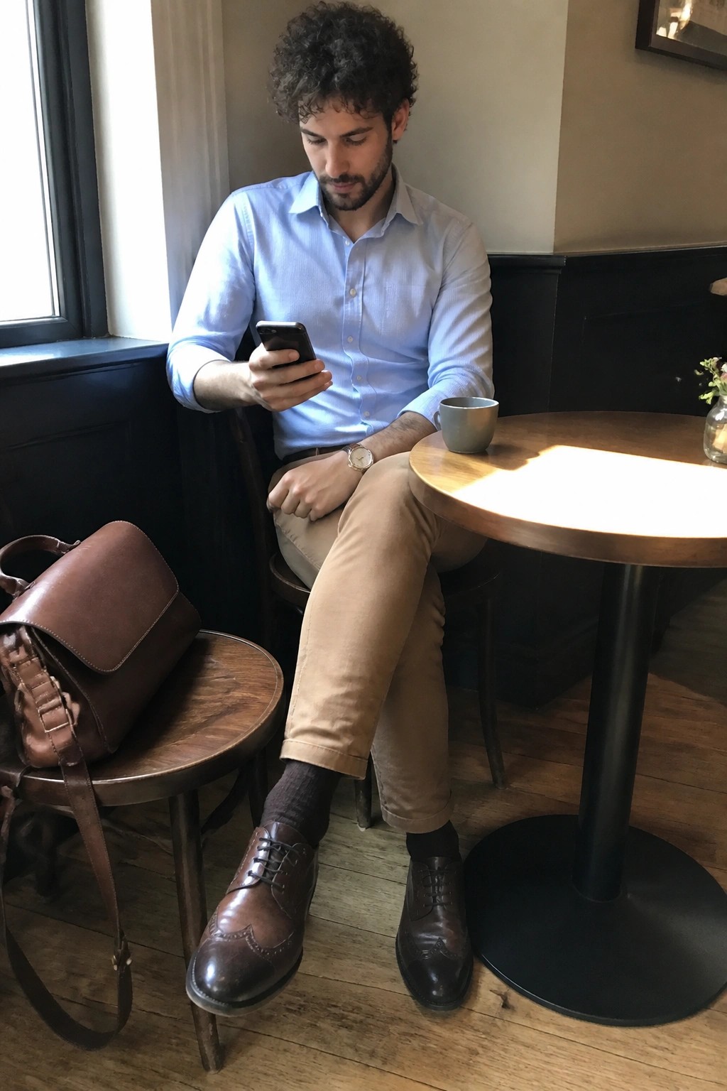 Man in blue shirt checks phone beside brown leather bag at wooden cafe table.