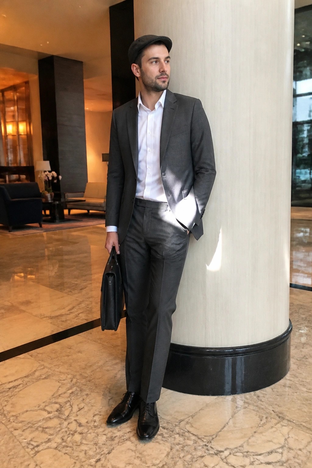 Man in gray suit and flat cap holds briefcase in marble lobby.