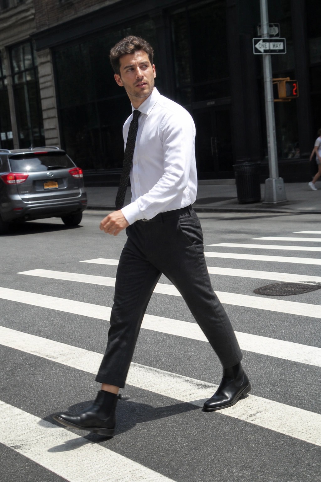 Man wearing a white button down shirt black tie black formal trousers and black chelsea boots while crossing a city street.