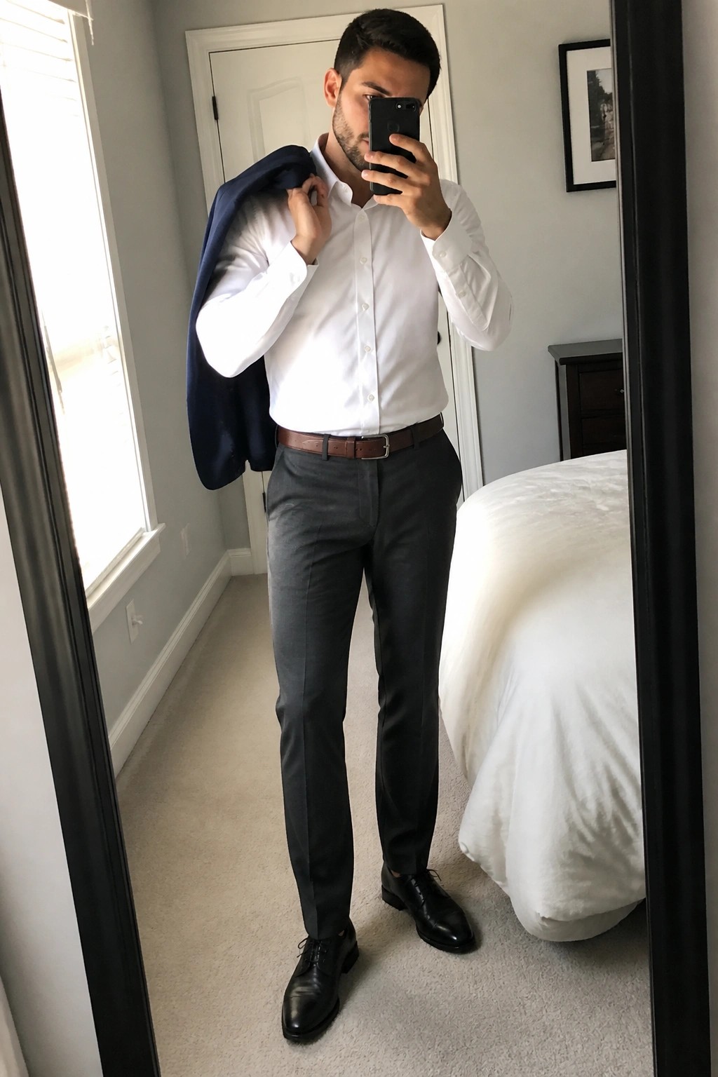 A man in a white dress shirt, gray formal trousers, brown belt, black dress shoes, holding a navy blazer over his shoulder while taking a mirror selfie.