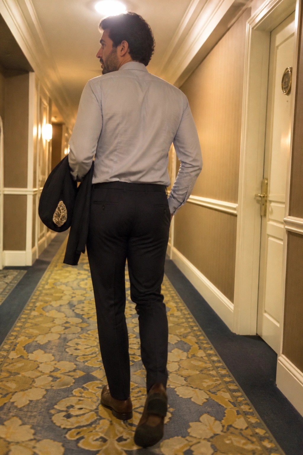 Man in blue shirt and dark pants walks patterned hallway carrying jacket over shoulder