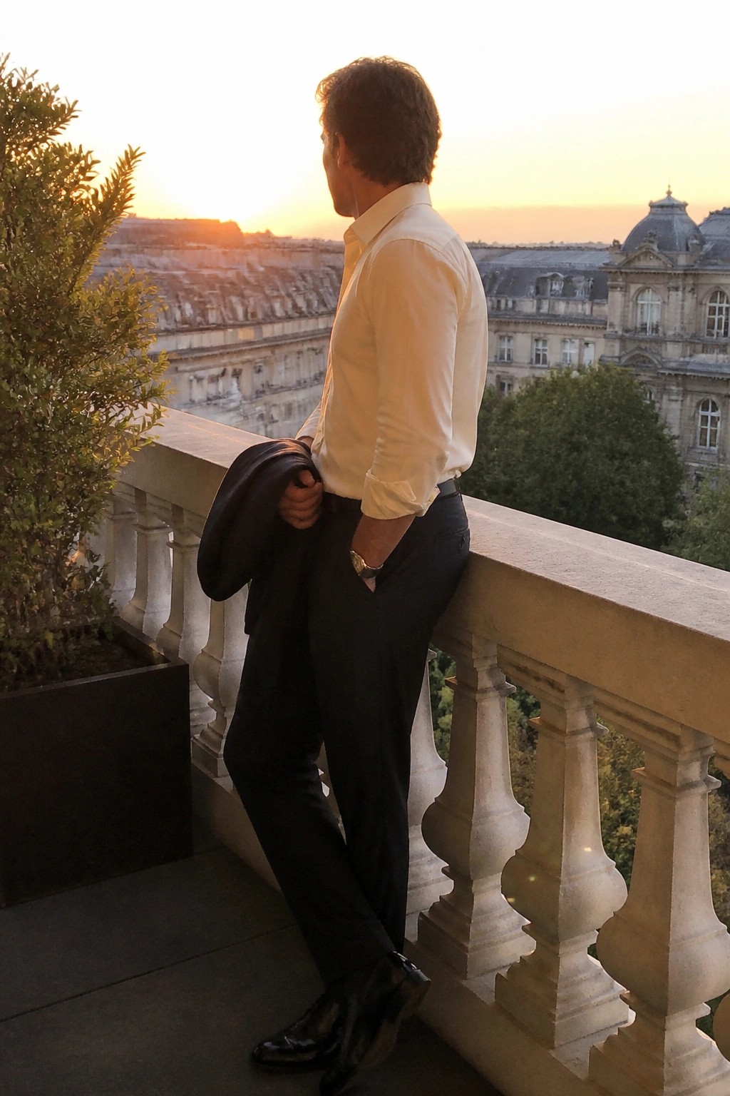Man wearing dark formal trousers with a white button down shirt, sleeves rolled, holding a matching jacket over one arm and black dress shoes visible on a balcony.