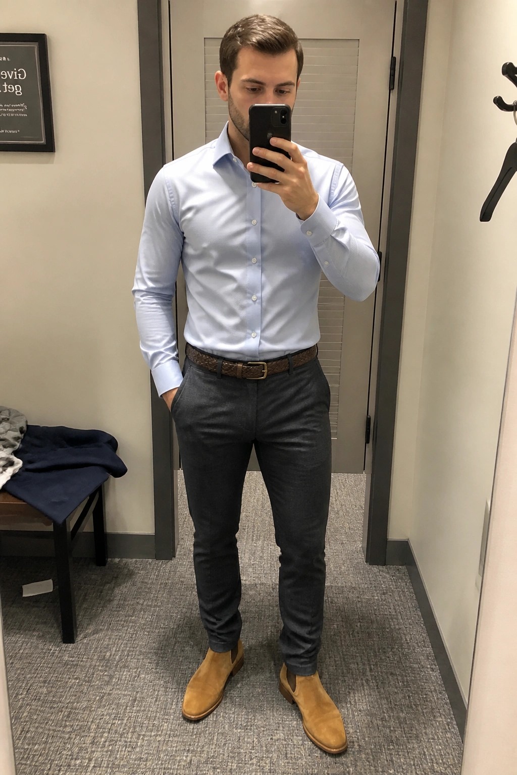 Man in light blue shirt and gray pants taking mirror selfie in dressing room