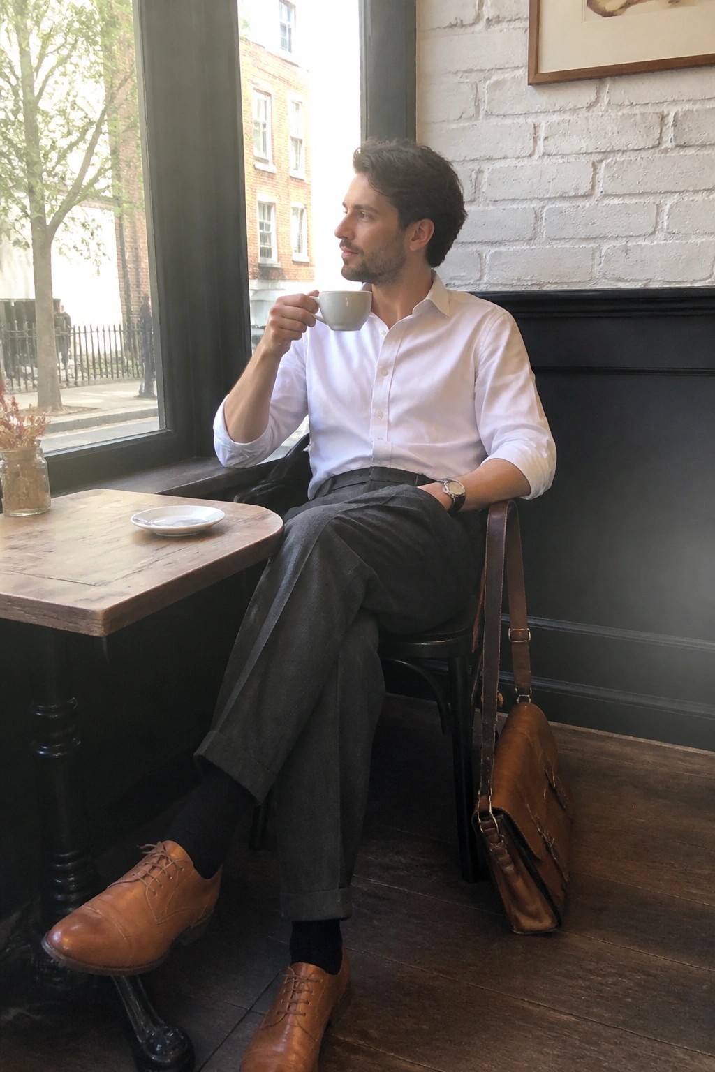 Man in white shirt sips coffee by cafe window with brown leather bag.
