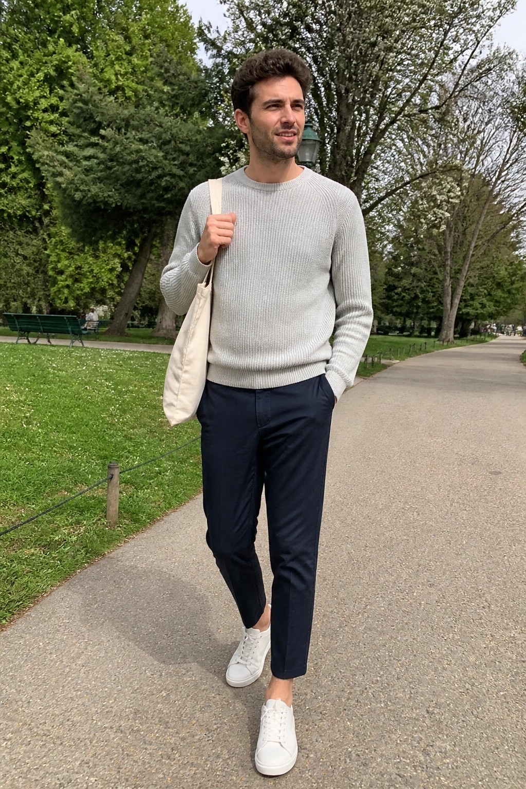 Man in light gray sweater and navy pants carrying beige tote in park