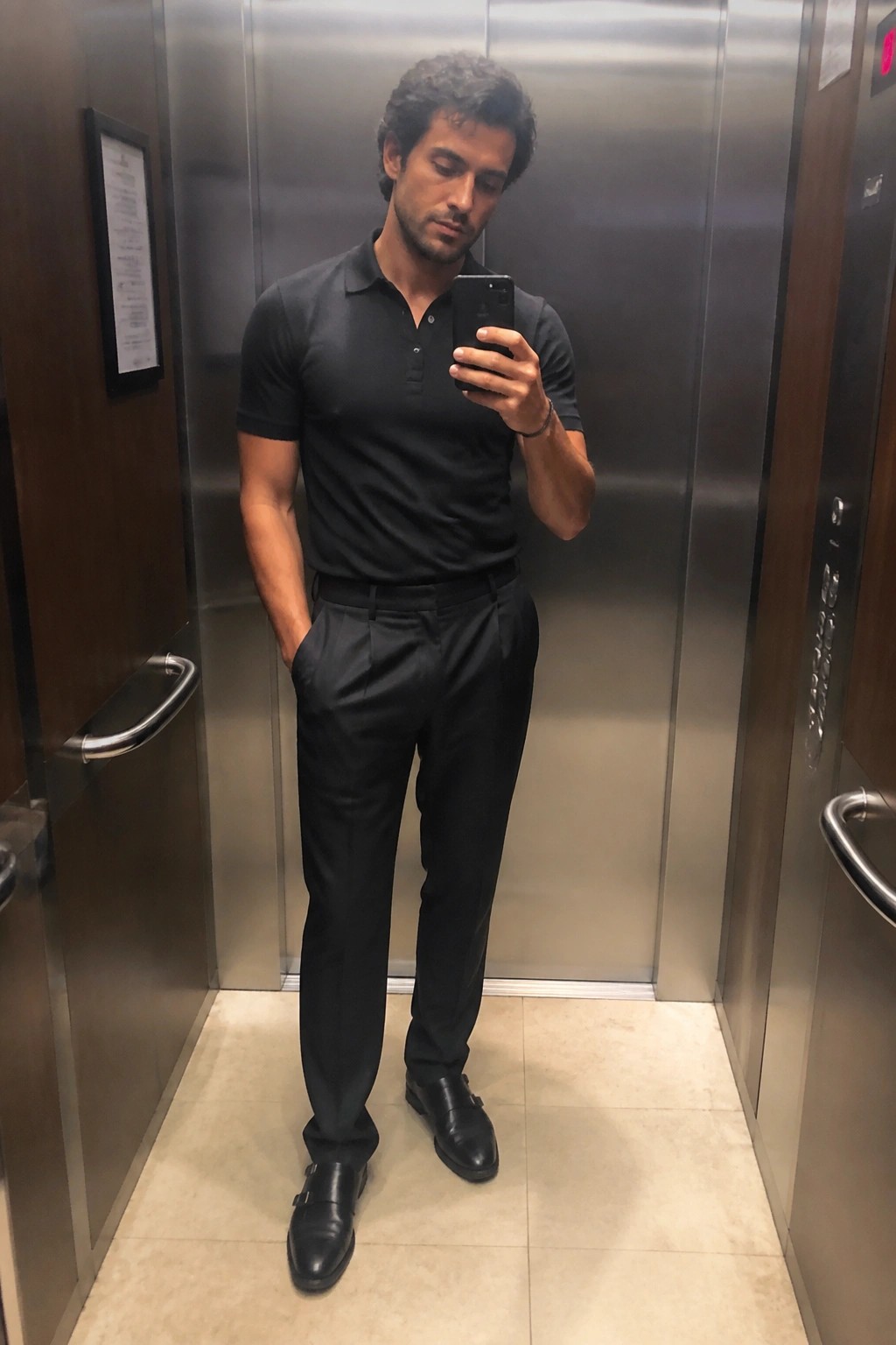 Man in black polo shirt and trousers taking selfie in elevator.