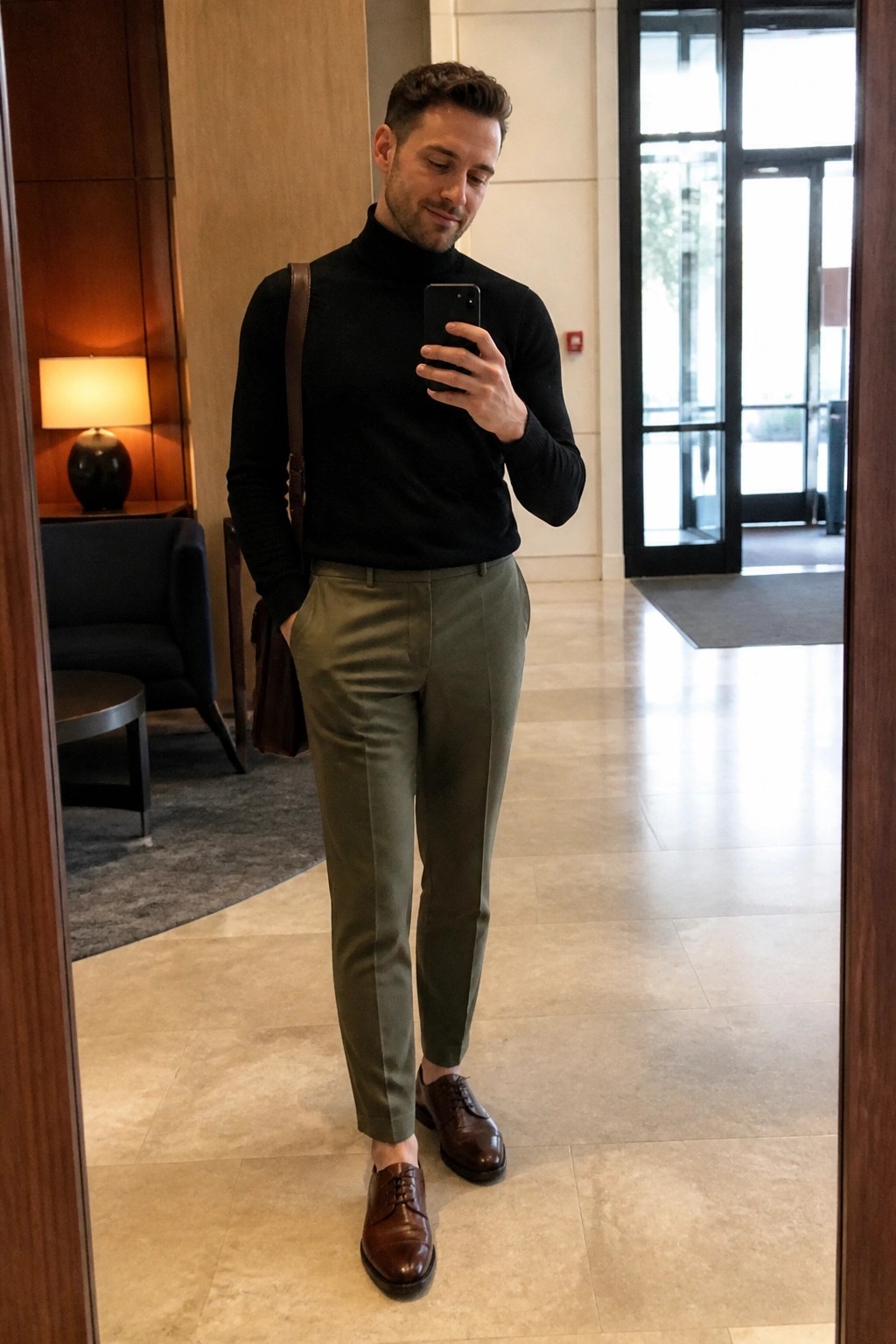 Man in black turtleneck and olive pants taking mirror selfie with phone.