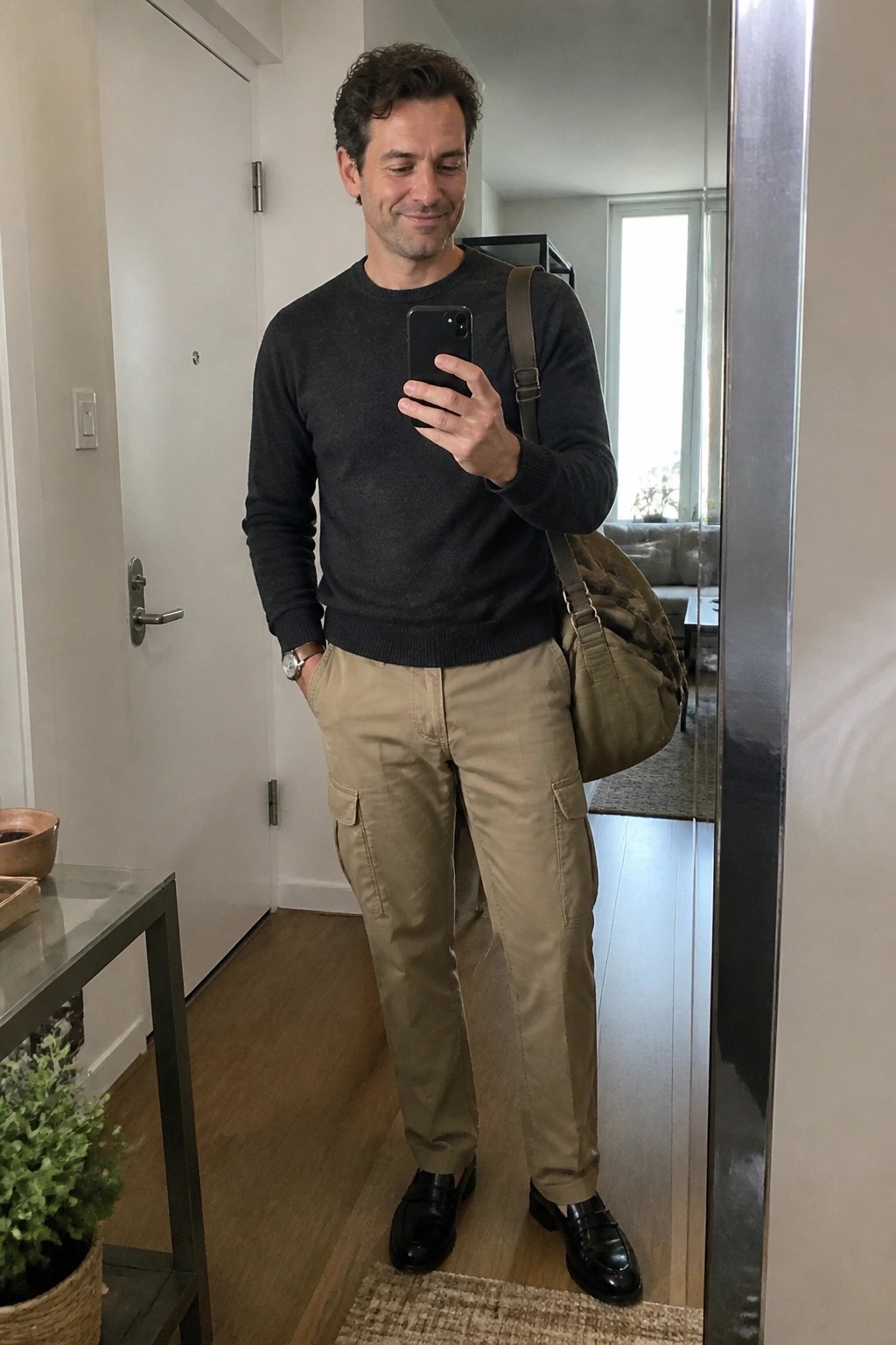Man in gray sweater and tan cargo pants takes mirror selfie with bag.