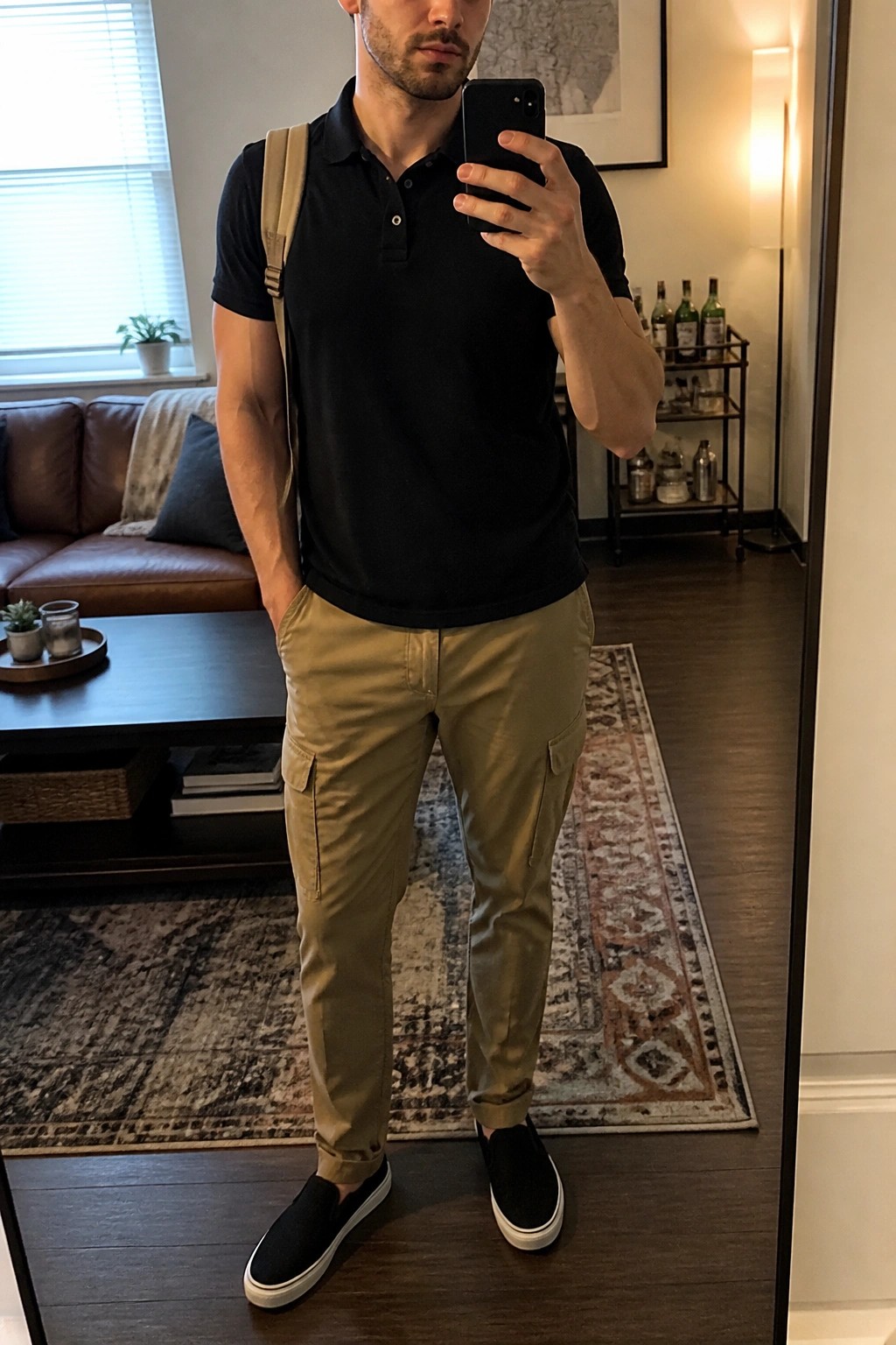 Man wearing black polo shirt tucked into khaki cargo pants with black slip-on shoes and beige backpack in a living room setting.