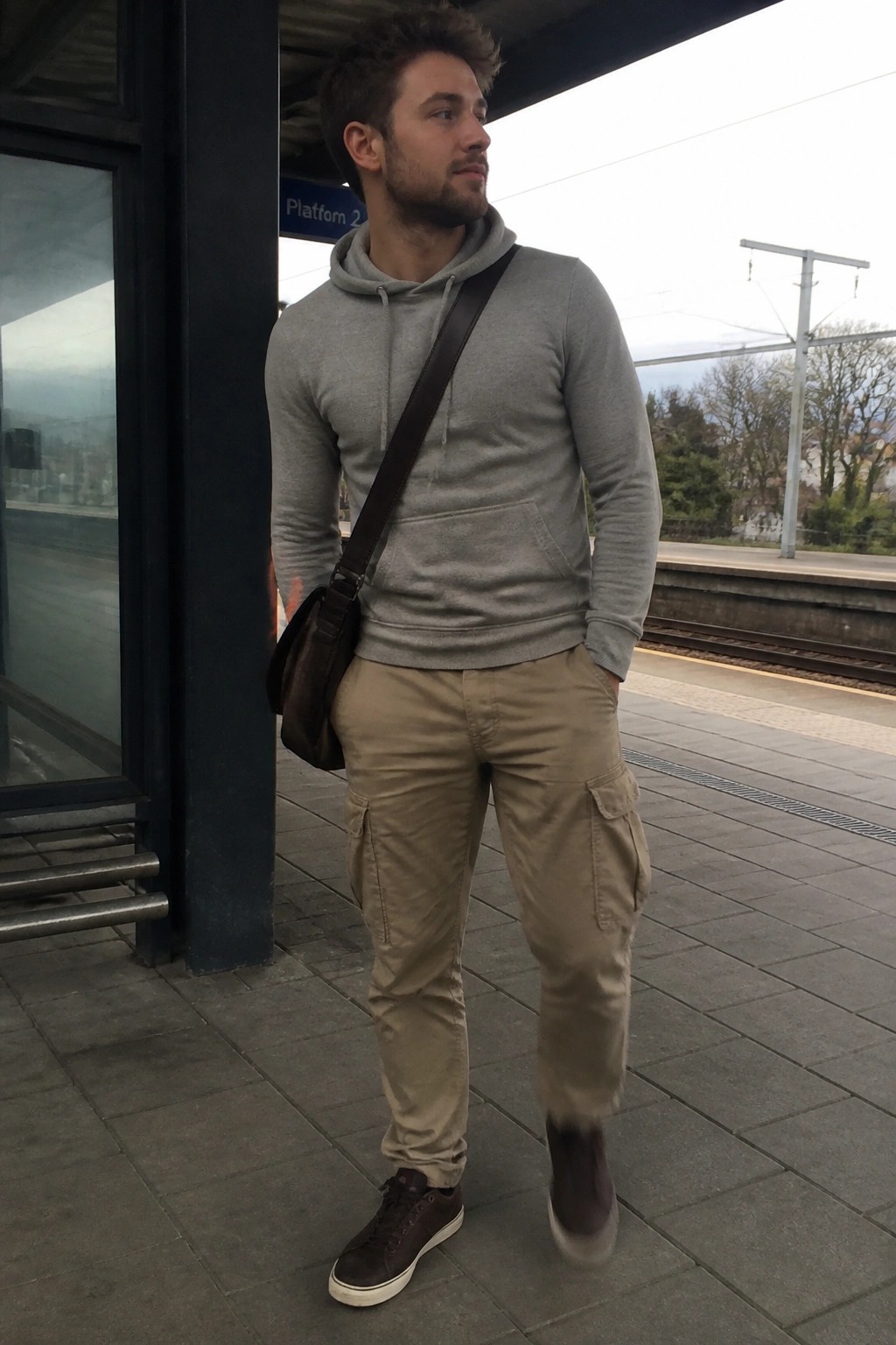 Young man in grey hoodie and cargo pants on train platform with shoulder bag