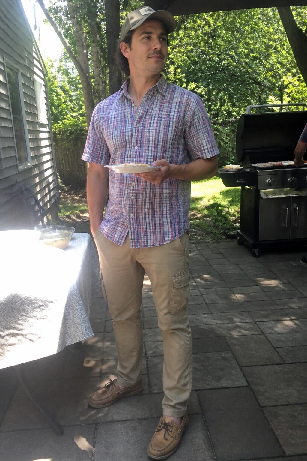 Man in plaid shirt and cap holds plate near grill on patio