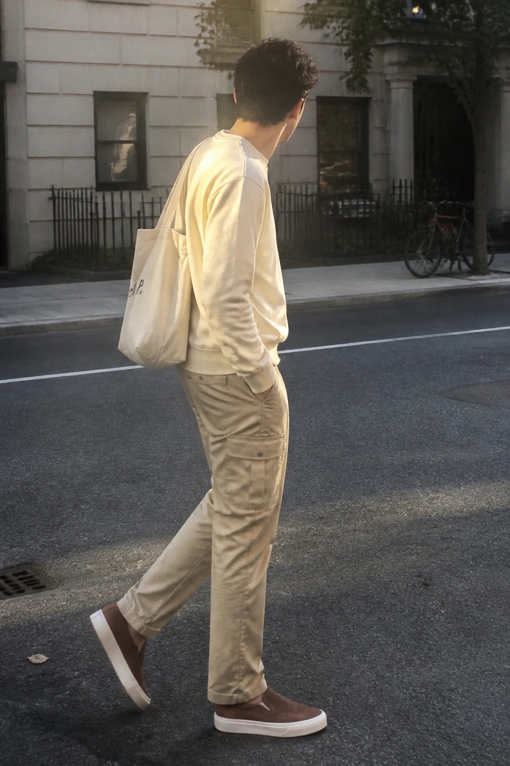Man in cream sweatshirt and beige cargo pants carrying tote bag on city street.