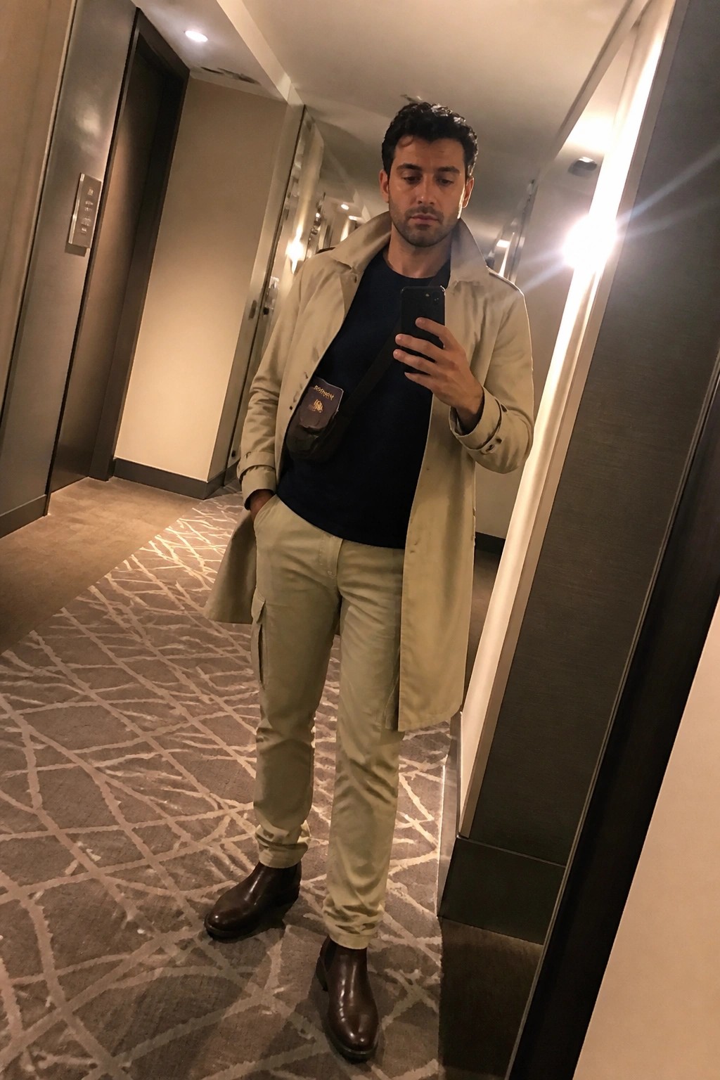 Man in beige trench coat and cargo pants taking mirror selfie in hallway
