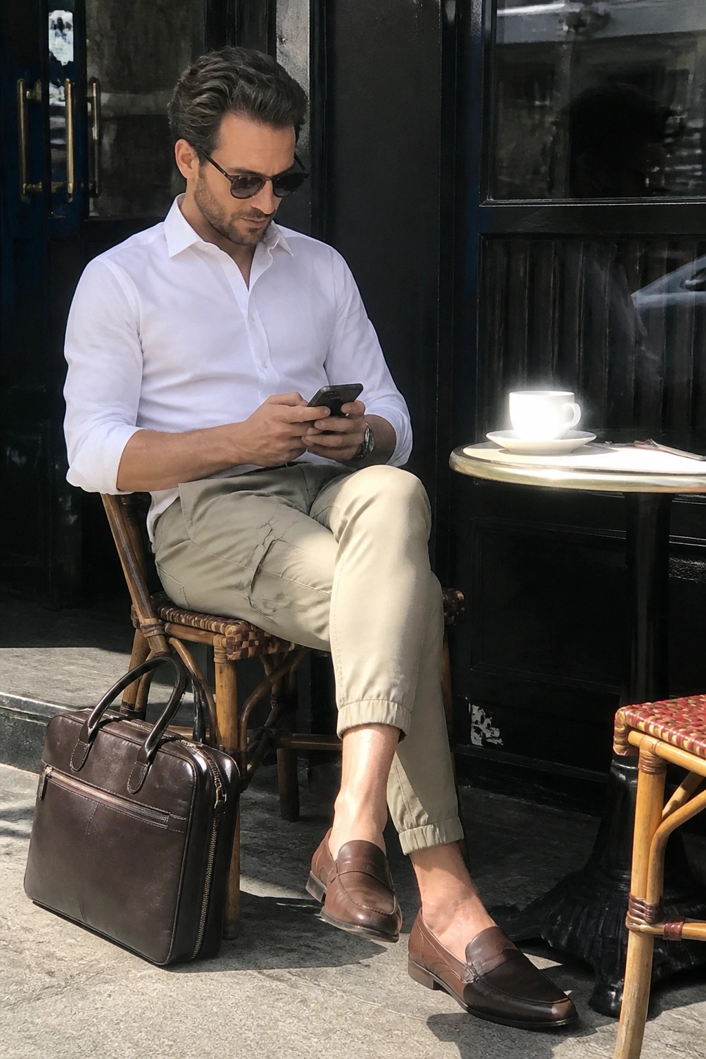 Man in white shirt checks phone beside brown briefcase at outdoor cafe.