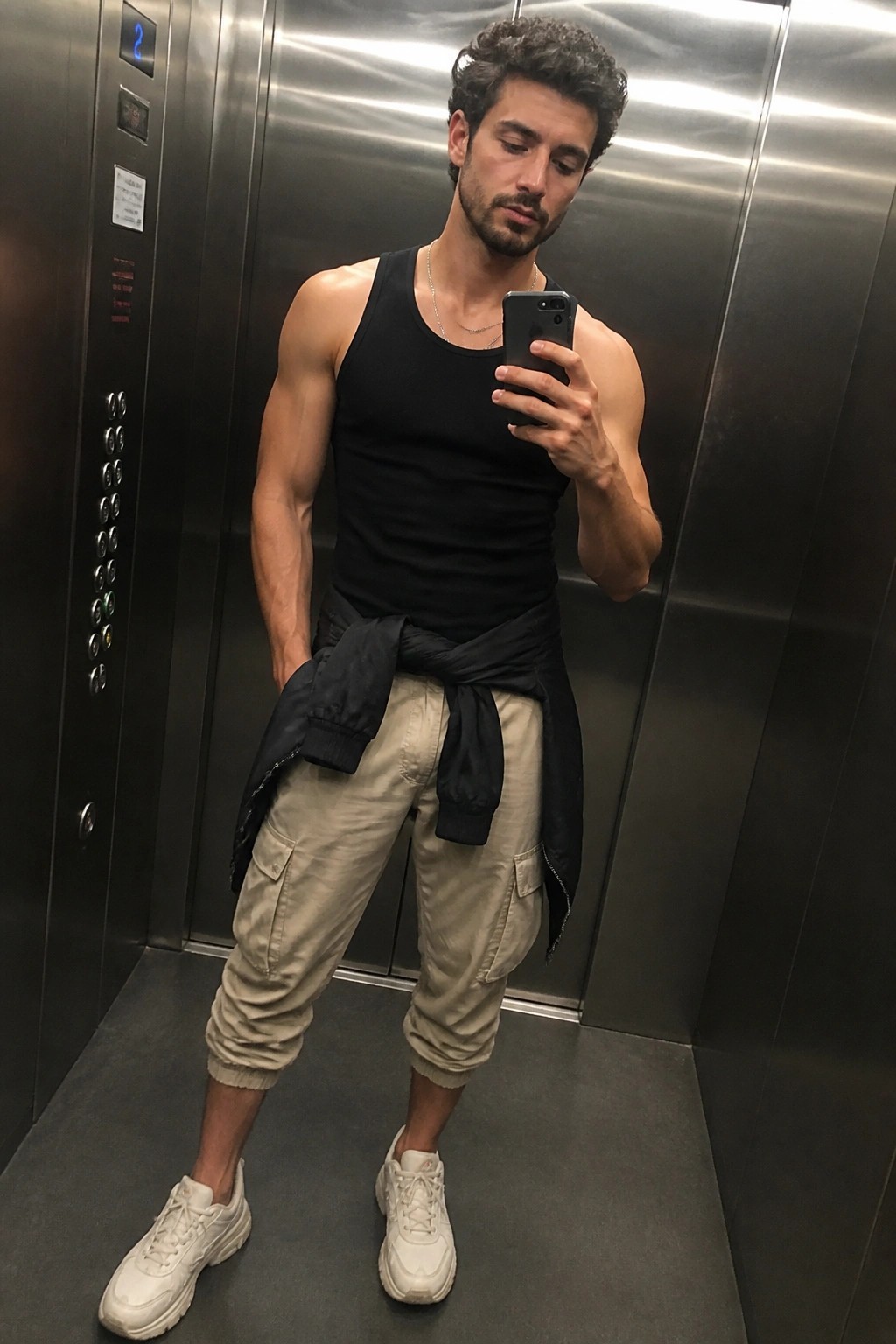 Muscular man in black tank top and beige cargo pants taking elevator selfie