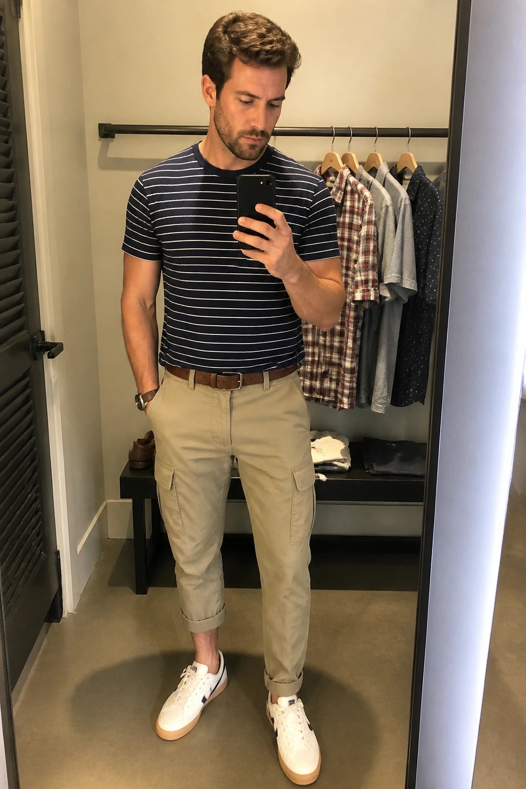 Man wearing navy striped short sleeve tee, khaki cargo pants with rolled cuffs, brown belt, and white sneakers while holding a phone in a closet setting.