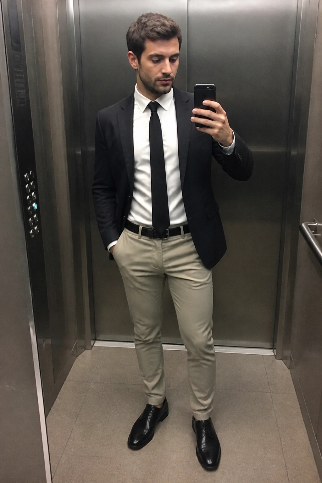 Man in black blazer, white shirt, black tie, beige pants taking elevator selfie