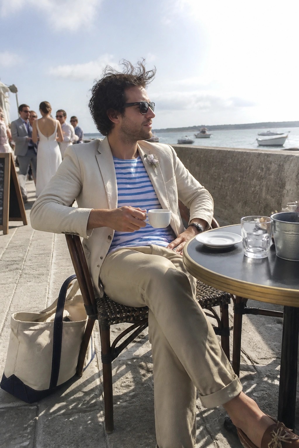 Man in striped shirt and blazer holds coffee at seaside table.